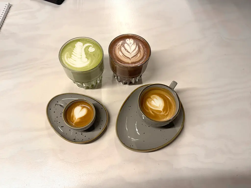 Das Bild zeigt vier verschiedene Kaffee-Spezialitäten, jeweils mit verzierter Crema. Darunter Cappuccino, Espresso und Matcha-Latte