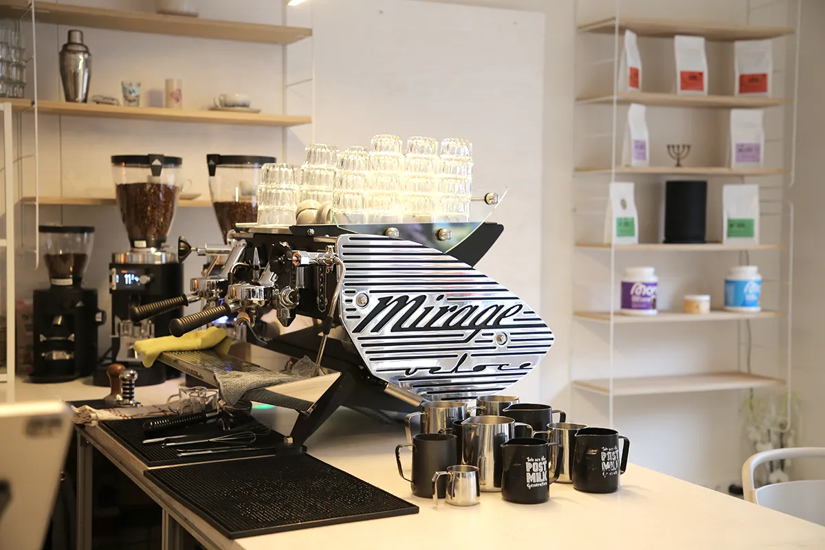 Das Bild zeigt eine große Gastronomie-Siebträger-Kaffeemaschine der Marke Mirage auf einem Tresen. Im Vordergrund stehen Tassen und Geschirr zur Kaffee-Zubereitung. Im Hintergrund zwei Kaffeemühlen, gefüllt mit Kaffeebohnen.